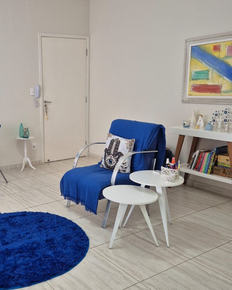 Consultório de Aline Calaes com poltrona azul, mesinhas brancas, estante de livros e quadro colorido