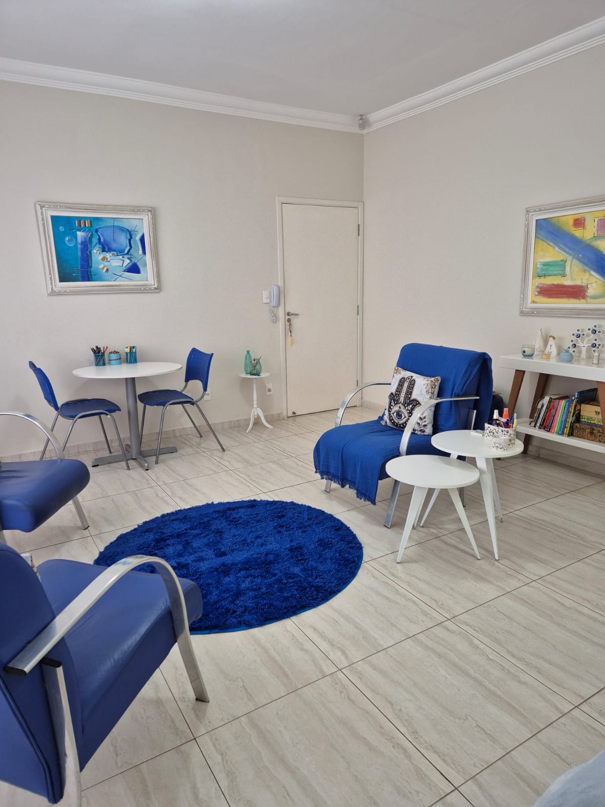 Consultório com quadros coloridos, mesa de atividades, poltronas azuis e estante de livros